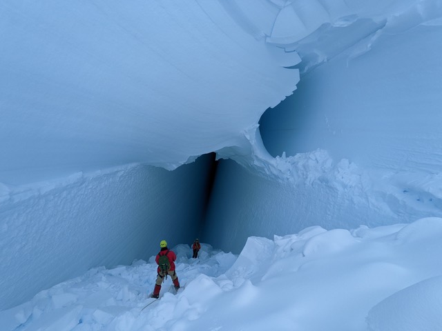 2 people stand on a ramp going down into a deep ice crevasse